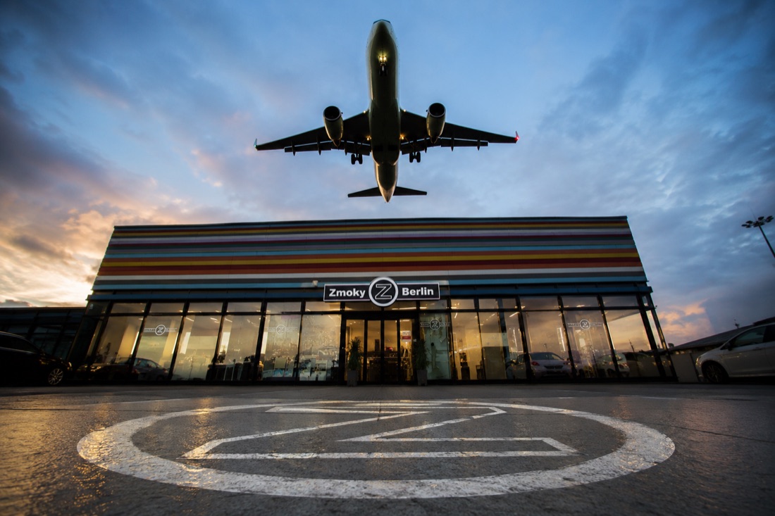Illustration: eine Zmokyz-Filiale am Stadtrand mit einem Flugzeug am Himmel